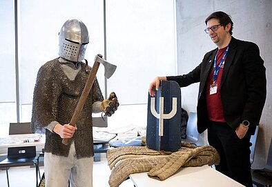 Persona viduslaiku bruņās un ķiverē tur kaujas cirvi, blakus stāv cilvēks ar vairogu un informācijas materiāliem uz galda