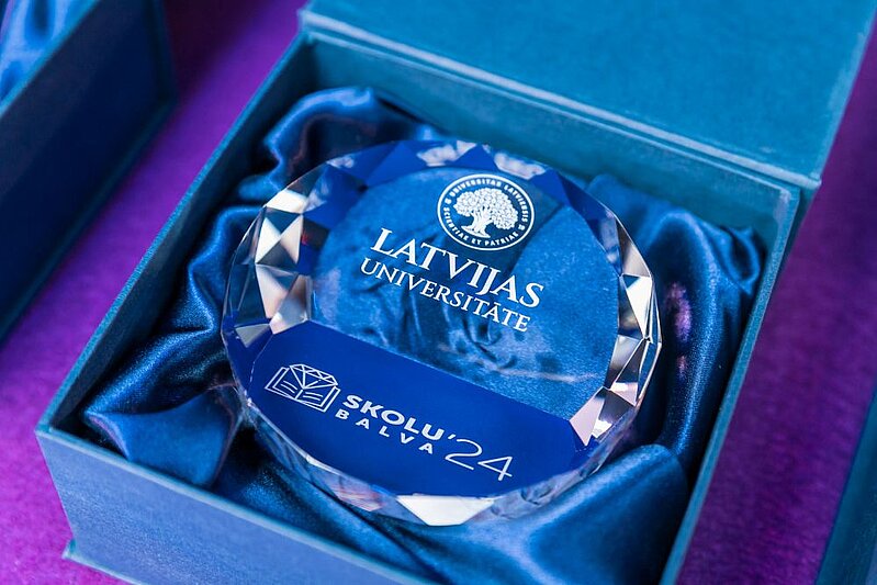 Latvijas Universitātes kristāla balva ar uzrakstiem ‘Latvijas Universitāte’ un ‘Skolu balva ’24’, novietota atvērtā kastē ar zilu satīna oderējumu.