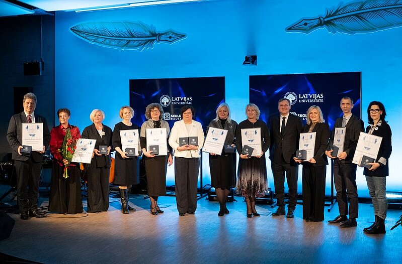 Grupa cilvēku stāv rindā uz skatuves, turot rokās balvas un apliecinājuma rakstus Latvijas Universitātes apbalvošanas pasākumā