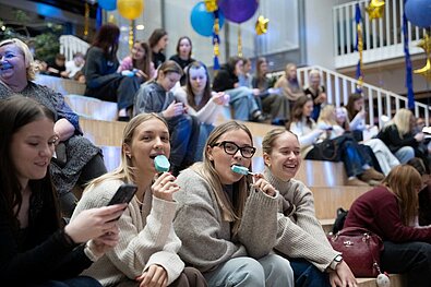 Jaunieši sēž uz kāpnēm pasākuma norises vietā, apkārt redzami baloni un cilvēki, kas sarunājas un izmanto telefonu