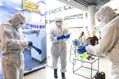 Trīs cilvēki ģērbušies baltos aizsargtērpos ar zilām cimdiem, gatavojas eksperimentam. Fonā redzams aprīkojums un uzraksts “Passion drives progress”.