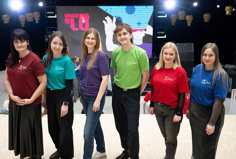 Seši cilvēki ar T-krekliem fakultāšu krāsās stāv rindā uz skatuves, aiz viņiem redzams liels ekrāns ar pasākuma vizuālo identitāti