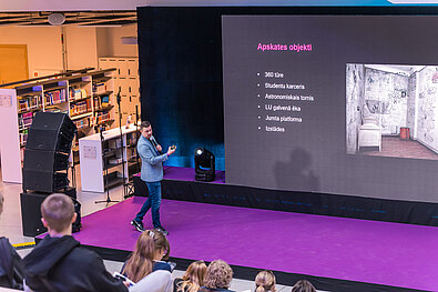 Cilvēks stāv uz skatuves ar mikrofonu rokā, prezentējot slaidu ar nosaukumu “Apskates objekti”. Uz ekrāna redzams saraksts: “360 tūre, Studentu karceris, Astronomiskais tornis, LU galvenā ēka, Jumta platforma, Izstādes” un fotogrāfija ar istabu, kuras sienas klātas ar grafiti. Fonā redzama bibliotēkas zona ar grāmatplauktiem.