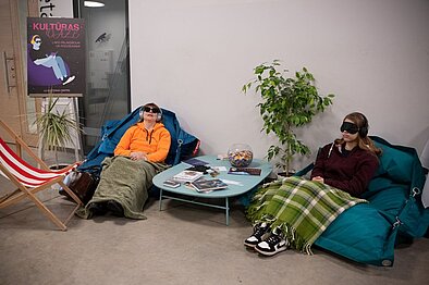 Divi cilvēki atpūšas sēžammaisos pie galdiņa ar žurnāliem un konfekšu trauku, pāri kājām pārklāti pledi, fonā plakāts un telpaugs