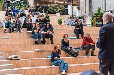 Cilvēki sēž uz plašām koka pakāpēm auditorijā, daži skatās telefonos, citi sarunājas. Fonā redzami augi un uzraksts “Radio TEV”.