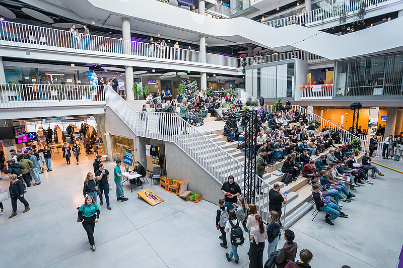 Liels, daudzlīmeņu iekštelpu ātrijs ar plaši apmeklētu pasākumu; cilvēki iet, pulcējas un sēž uz kāpnēm, bet augšējos stāvos redzami papildus skatītāji un aktivitātes.