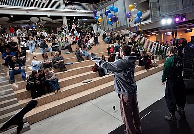 Skatītāji sēž plašā auditorijā ar kāpnēm, bet uz skatuves priekšā stāv mūziķis, pie griestiem un sānos redzami baloni un gaismas