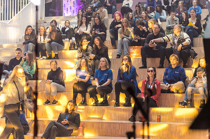 Plašas kāpņu tipa sēdvietas, kur cilvēki sēž, sarunājas un vēro notiekošo pasākumā; apgaismojums ir silts un atmosfērisks.