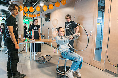Cilvēks sēž uz krēsla un tur metāla apli, demonstrējot fizikas eksperimentu. Apkārt stāv citi cilvēki, fonā pie sienas piestiprināti oranži baloni un galds ar aprīkojumu.