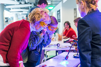 Cilvēki ar dzeltenām aizsargbrillēm skatās uz galdu, kur novietots mikroskops un apgaismots objekts ar zilganu gaismu. Fonā redzami citi cilvēki pie galdiem.