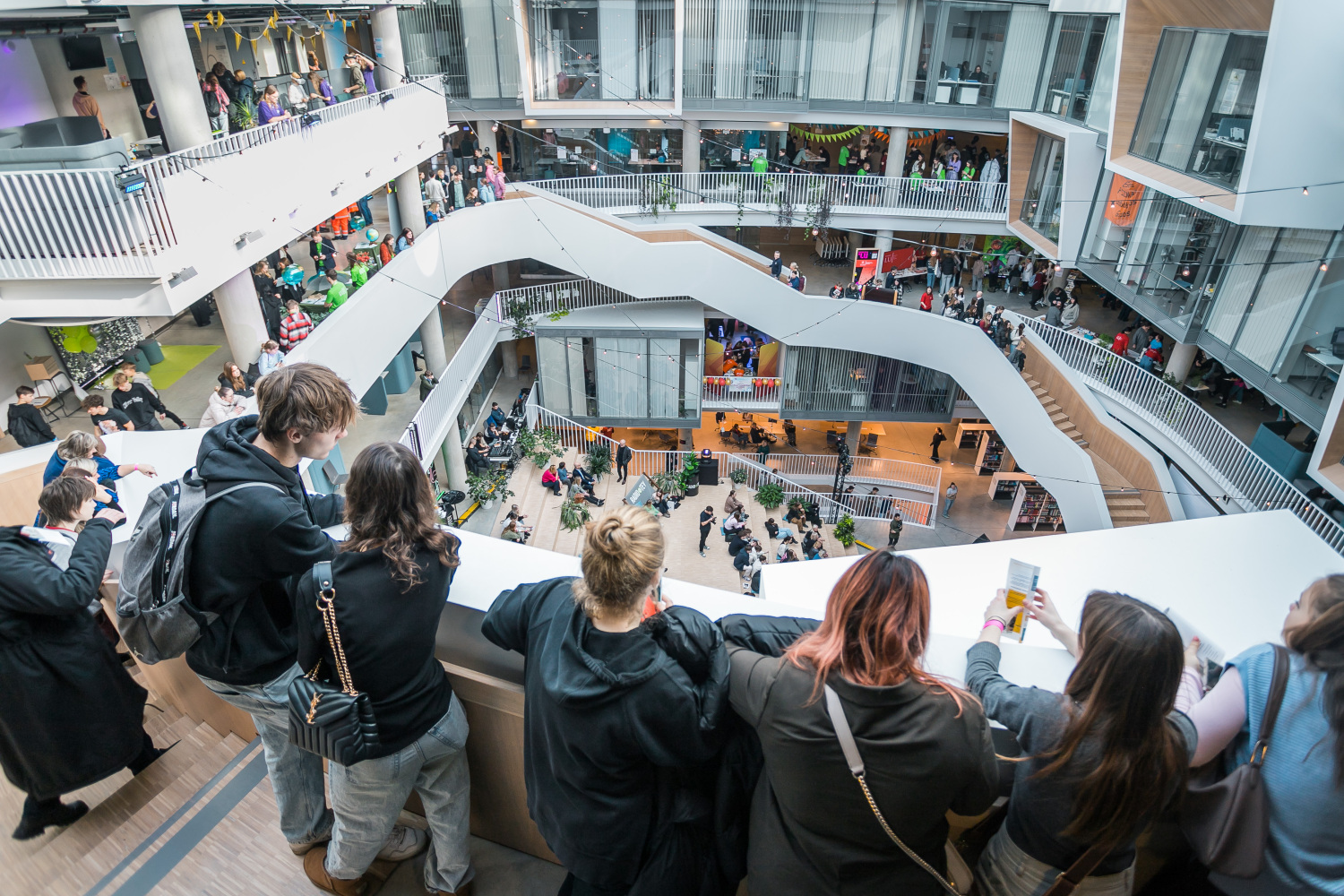 Vairāki jaunieši stāv augšējā stāvā LU Zinātņu mājā un noraugās uz norisēm zemāk