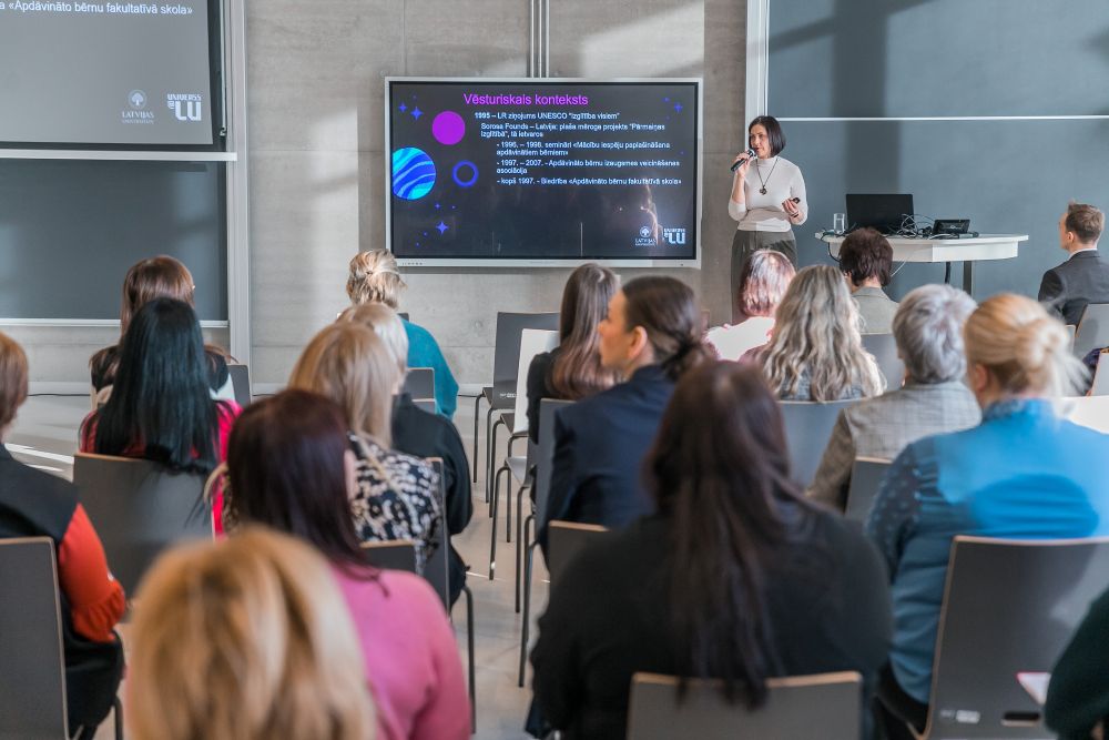 Lekcija auditorijā, kur cilvēki sēž rindās un klausās runātāju, kas stāv pie ekrāna un prezentē informāciju par visuma vēsturi; ekrānā redzams slaids ar tekstu un ilustratīviem planētu attēliem.
