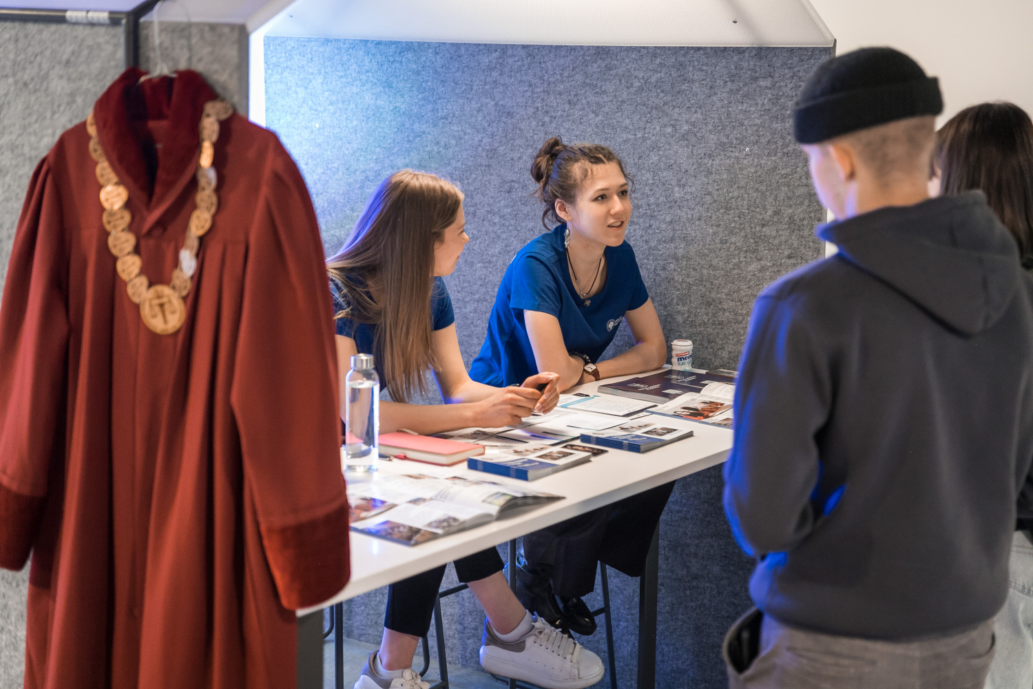 Informācijas galds pasākumā, kur divi cilvēki sēž pie augsta galda, sarunājas un izskaidro materiālus apmeklētājiem; priekšplānā redzams akadēmisks tērps ar ķēdi.