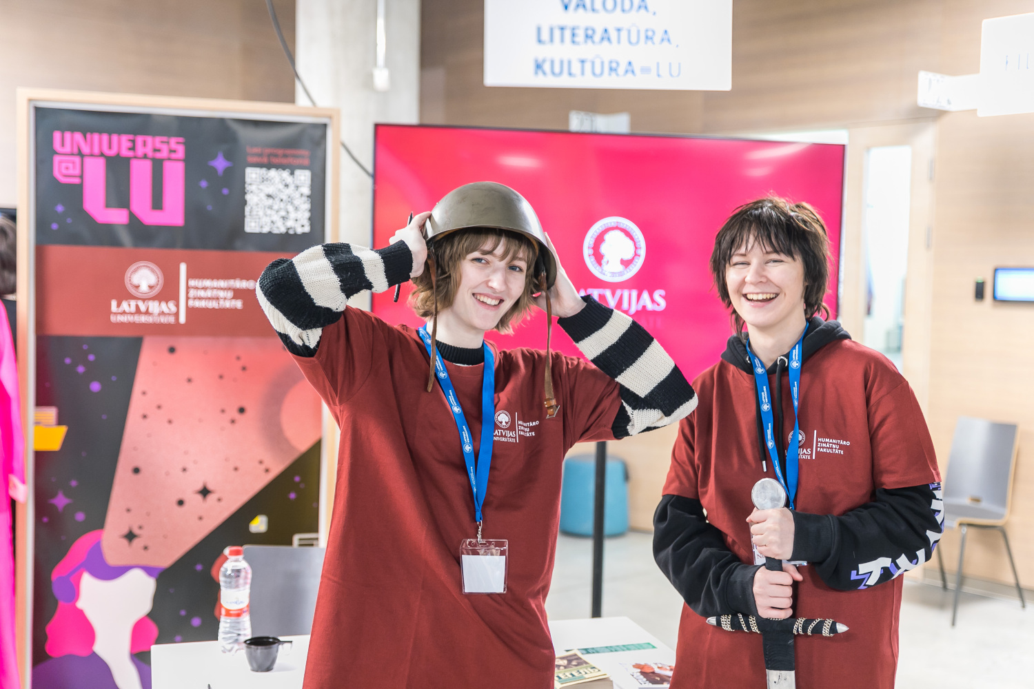 Divi jaunieši stāv pie Latvijas Universitātes informatīvā stenda; abi valkā sarkanus pasākuma krekliņus ar identifikācijas kartēm. Viens tur uz galvas militāra stila ķiveri, otrs rokās tur mikrofonu. Aiz viņiem redzams liels ekrāns ar Latvijas Universitātes simboliku un stenda materiāliem.